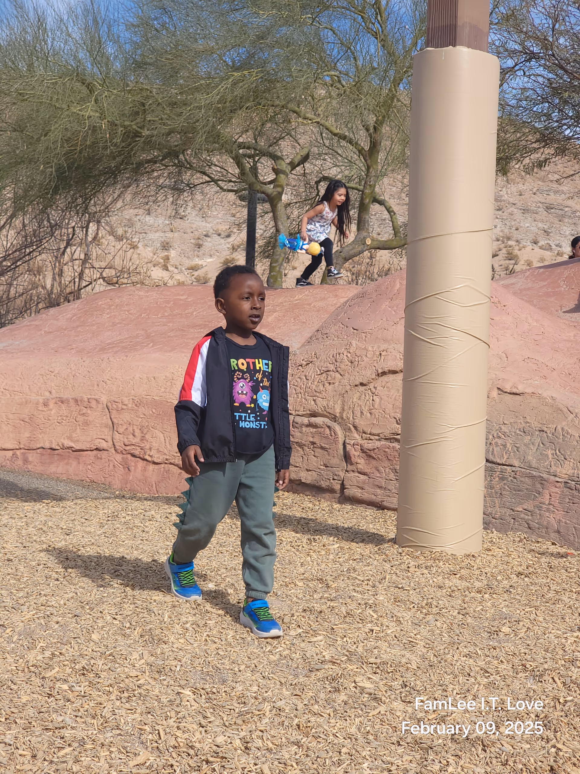A young child walks on wood-chip ground beside a padded outdoor play area.