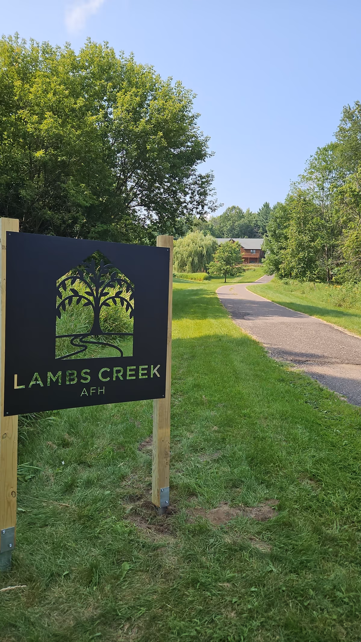 Sign for Lambs Creek AFH beside a paved path through a grassy, tree-lined campus.