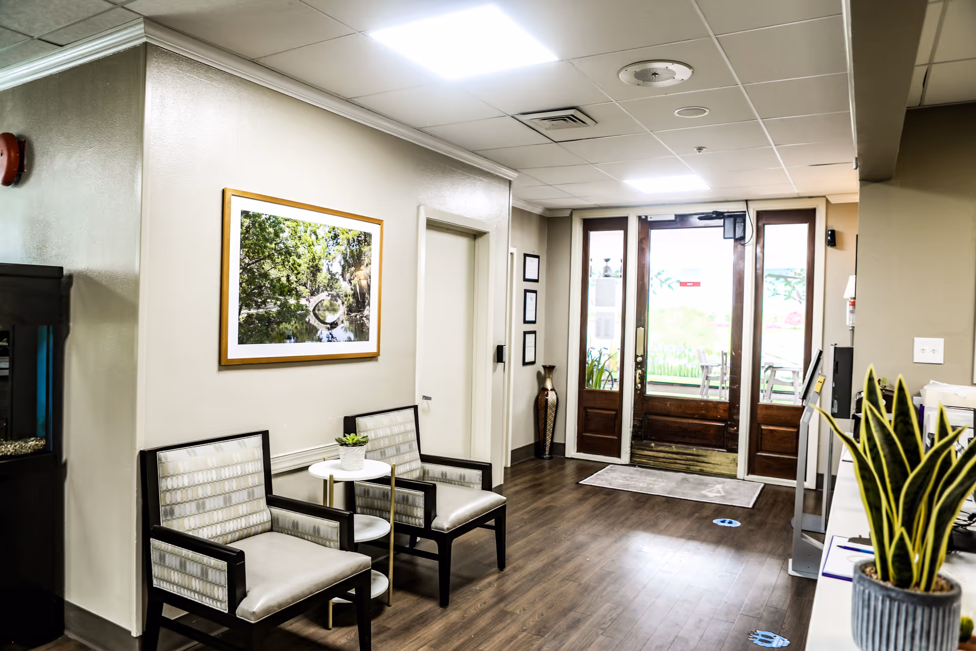 Bright senior living facility entryway with two armchairs, a small round table and plant, framed artwork on the wall, and double glass doors leading outside.