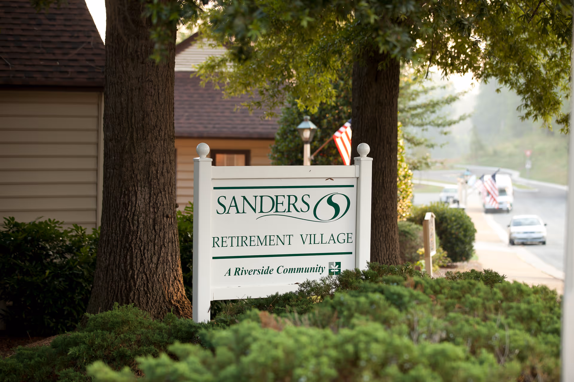 Sanders Retirement Village sign with trees and greenery beside a street.