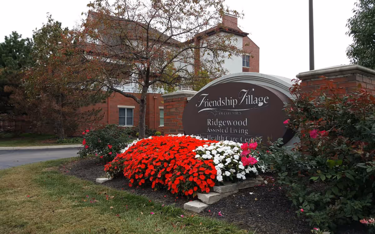 Outdoor view of a senior living facility entrance with a large sign reading 'Friendship Village of Columbus Ridgewood Assisted Living & The Health Care Center' surrounded by vibrant red, white, and pink flowers, with a brick building and trees in the background.