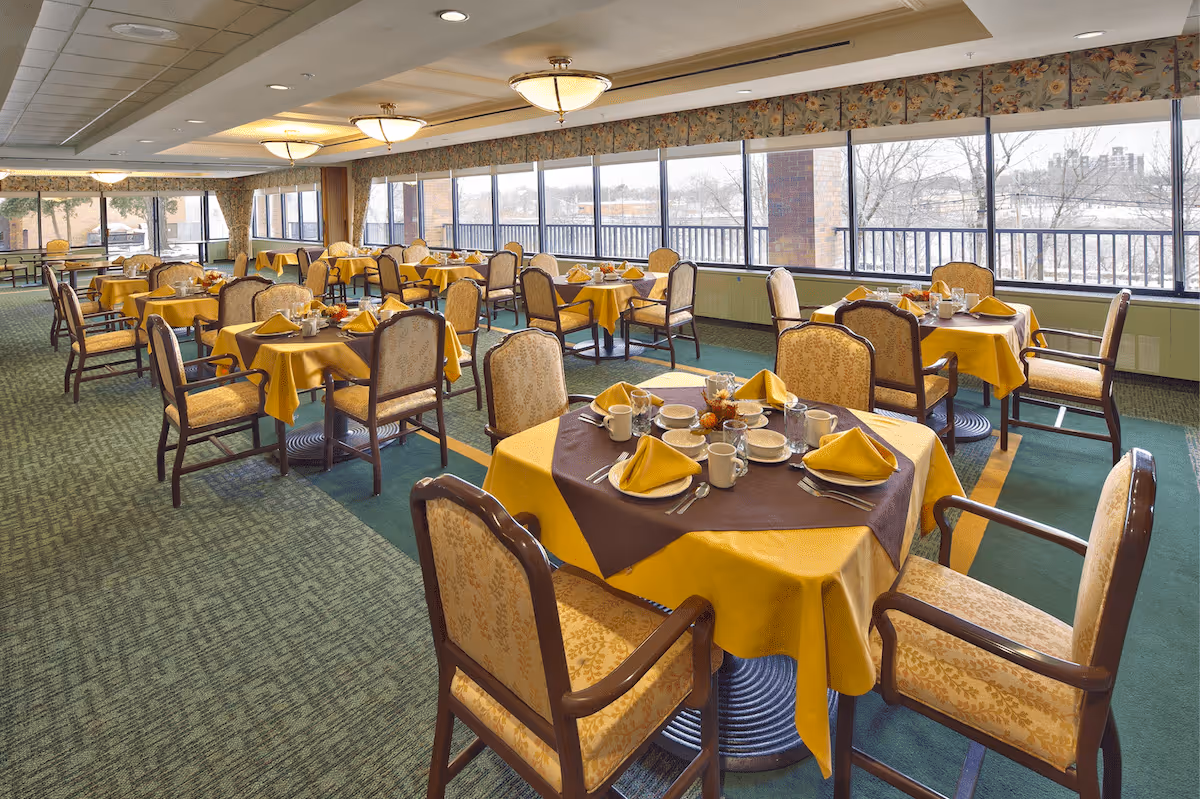 A spacious dining room with multiple tables covered in yellow tablecloths and brown overlays, each set with cups, plates, silverware, and folded yellow napkins. The room has large windows along one side, allowing natural light to fill the space and offering a view of the outdoors. The chairs have wooden frames with patterned upholstery, and the ceiling features round light fixtures.