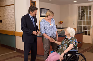 A woman in a wheelchair is shaking hands with a standing woman in a patterned blouse, while a man in a suit looks on in a hallway or reception area of a nursing and rehab facility.