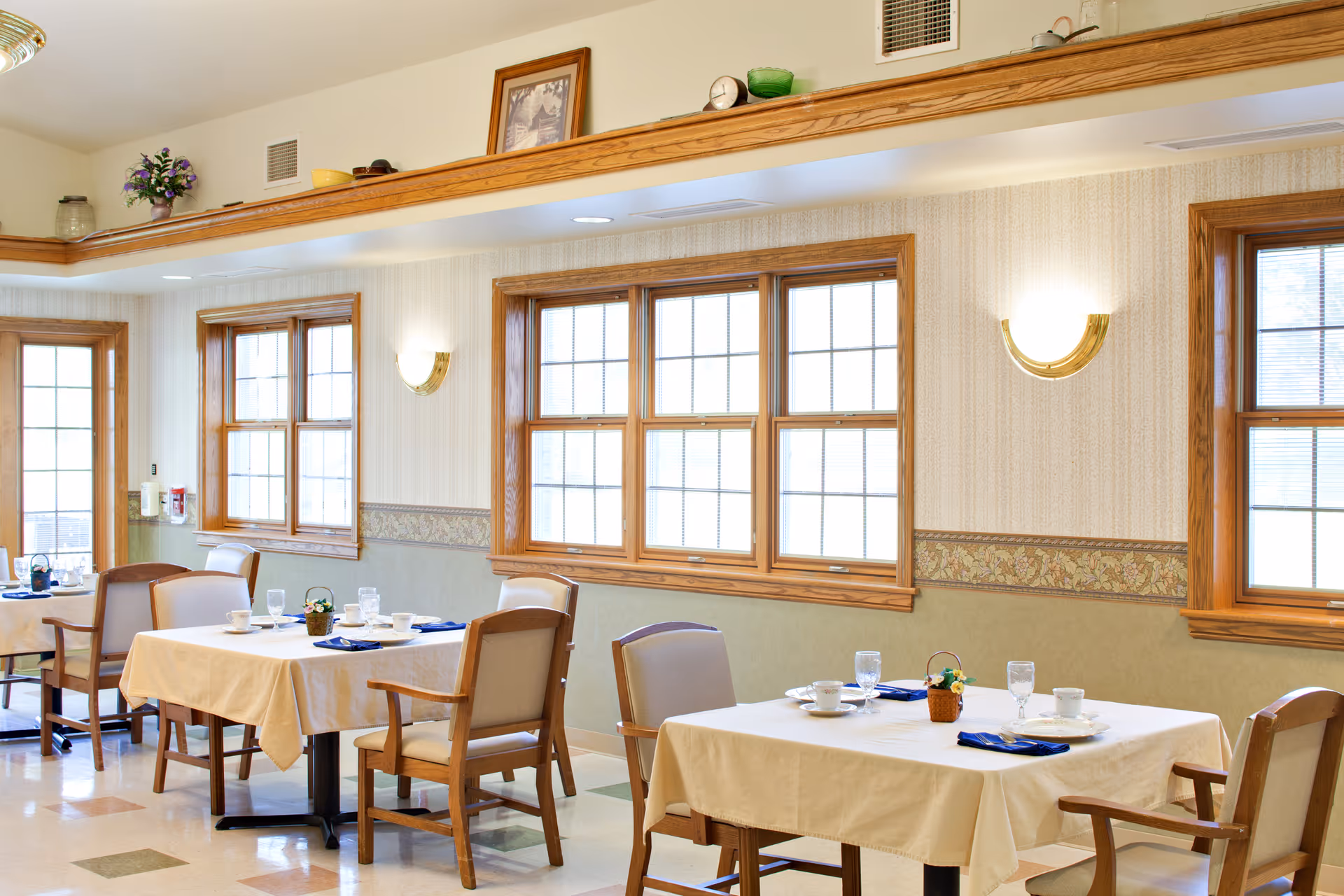 A bright dining room with several tables covered in beige tablecloths, each set with cups, glasses, plates, and blue napkins. Wooden chairs with cushioned seats surround the tables. The room has large windows with wooden frames, patterned wallpaper, and wall-mounted light fixtures. Decorative items are placed on a ledge near the ceiling.