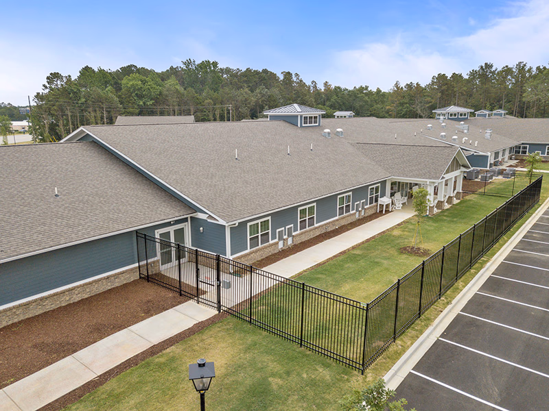Exterior view of a single-story senior living facility building with a gray roof and blue siding. The building is surrounded by a black metal fence enclosing a grassy area with a sidewalk. There is a parking lot adjacent to the building and trees in the background under a partly cloudy sky.