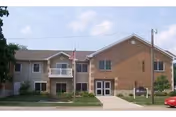 Two-story senior living building with brick facade, lawn, and American flag at entrance.