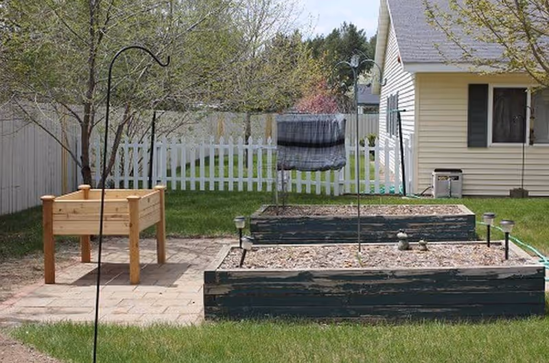 A backyard with raised garden beds, a wooden planter, a white fence, and a house.