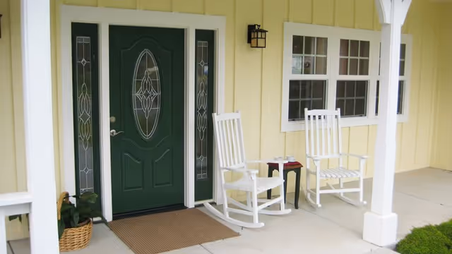 Front porch of a yellow house with a dark green door featuring decorative glass panels. Two white rocking chairs and a small black side table with a red book are placed on the porch. A basket with greenery is near the door, and there is a wall-mounted light fixture above the chairs.