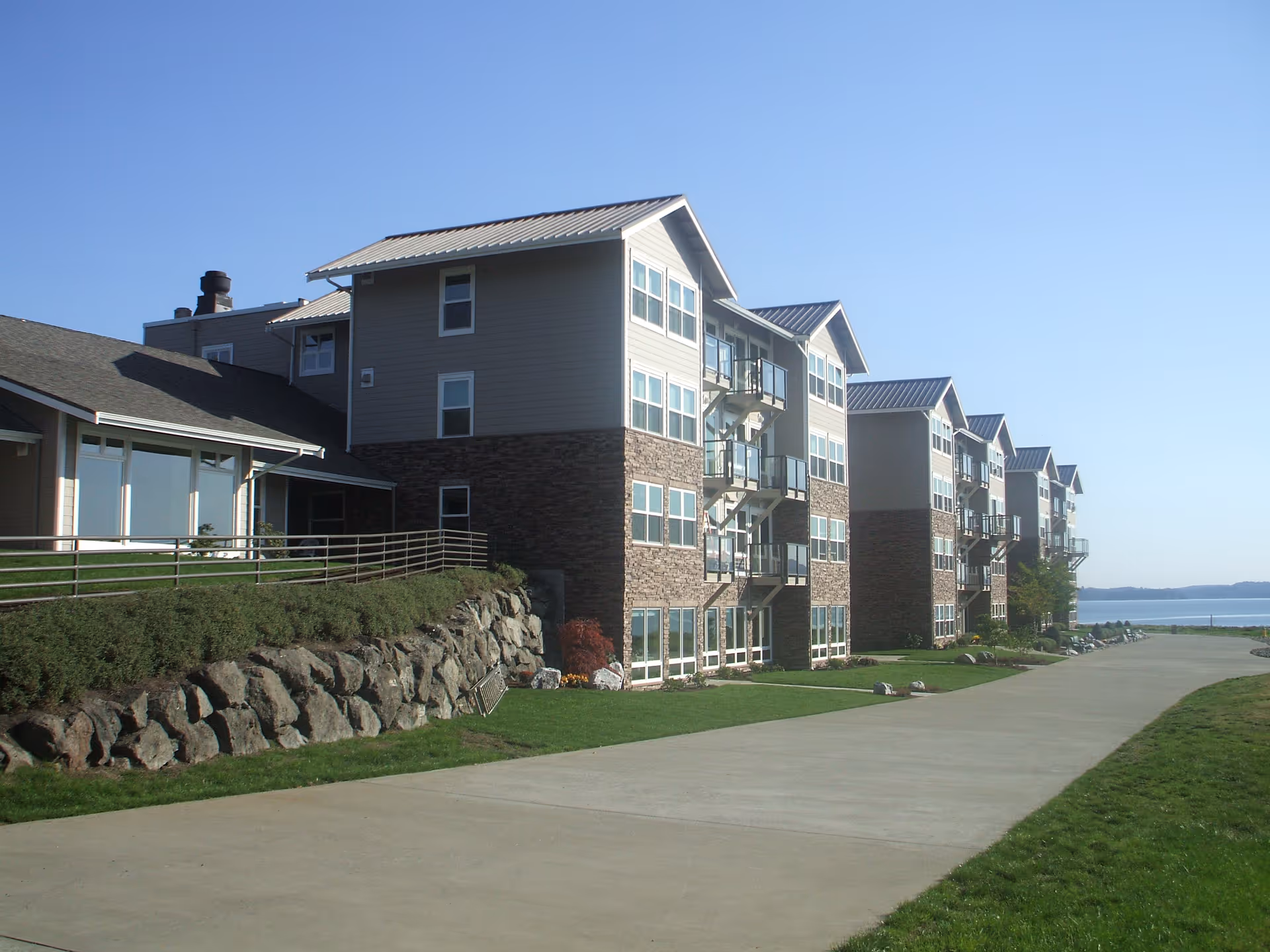 Multi-story waterfront senior living building with balconies, a paved walkway, and landscaped lawn.