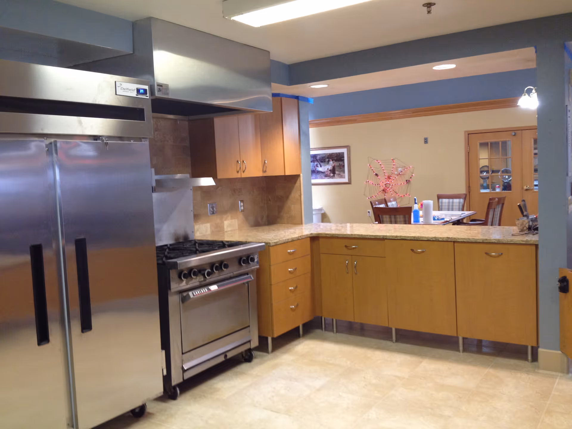A kitchen area with stainless steel double-door refrigerator and a commercial stove with oven. Light wood cabinets and drawers with a granite countertop form an L-shaped counter. In the background, there is a dining area with a table, chairs, and a decorative piece on the wall.