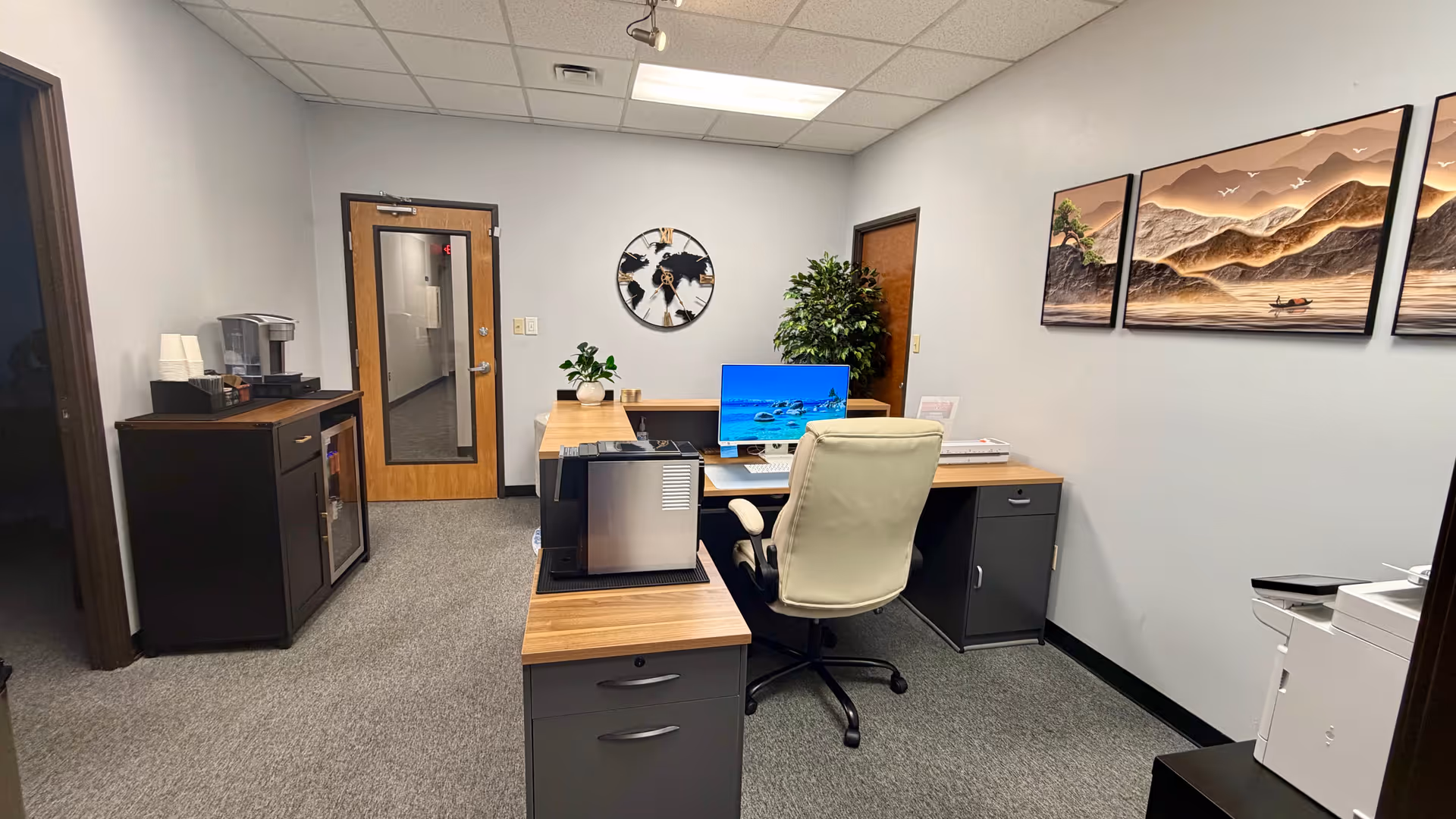 Office area with desks, a computer monitor, printer, coffee station, and wall art.