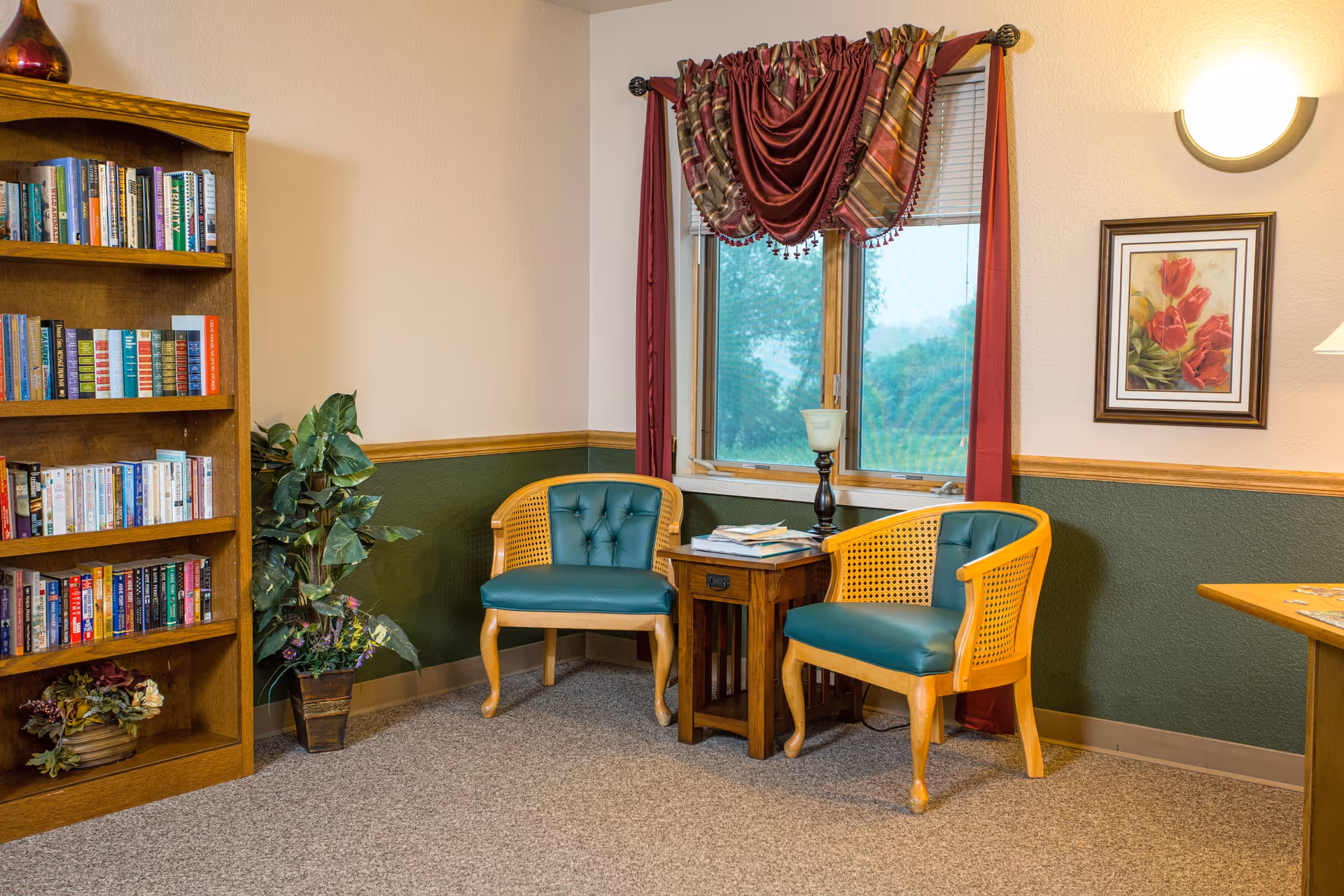 A cozy sitting area in a room with two wooden chairs with green cushions, a small wooden side table with books and a candle holder, a window with red and striped curtains, a bookshelf filled with books, a potted plant, a framed floral painting on the wall, and a wall-mounted light fixture.