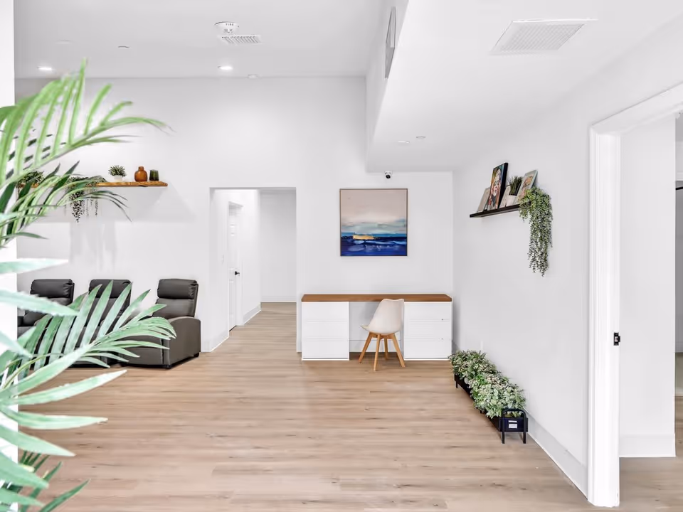 Bright open lounge area with a wood floor, a desk and chair against a white wall, reclining chairs, and potted plants.