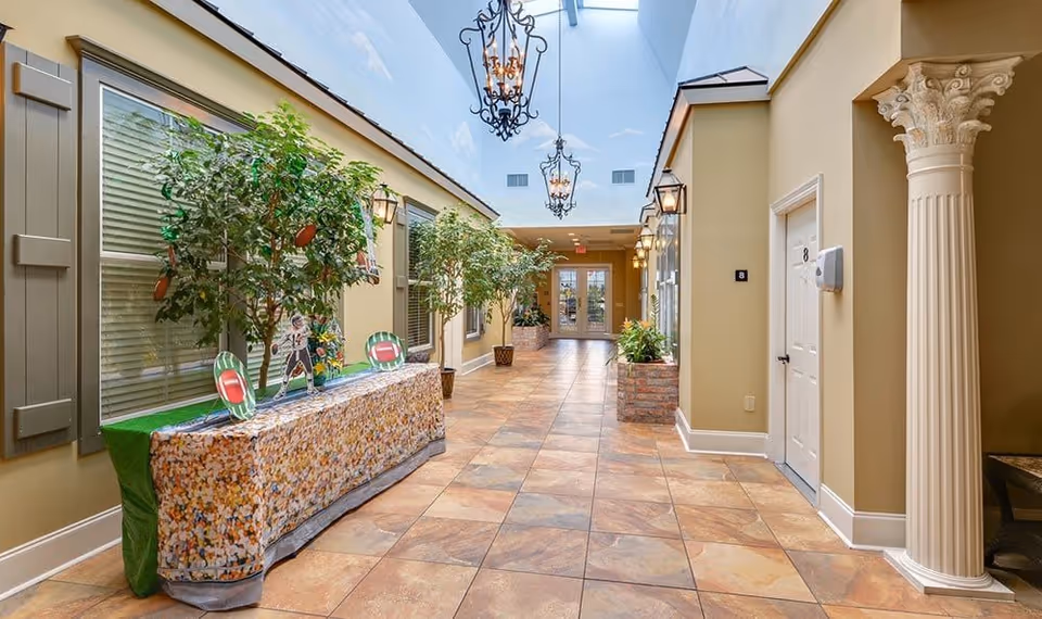 A bright indoor hallway with large windows on the left side, potted plants along the corridor, decorative hanging lanterns from the ceiling, and a white door labeled with the number 8 on the right. The floor is tiled with a warm-toned pattern, and there is a decorative column on the right side near the door.