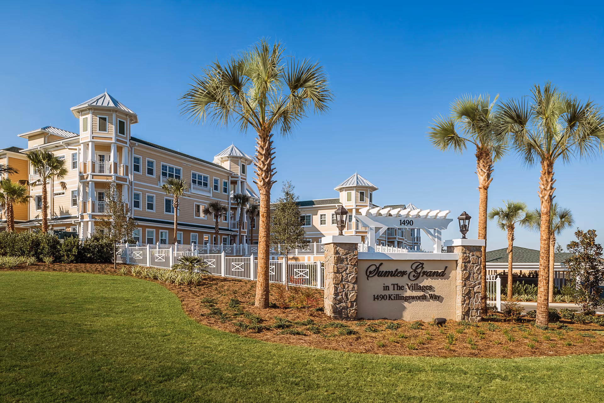 Exterior view of Sumter Grand senior living facility with a large sign displaying the name and address, surrounded by palm trees and well-maintained landscaping under a clear blue sky.