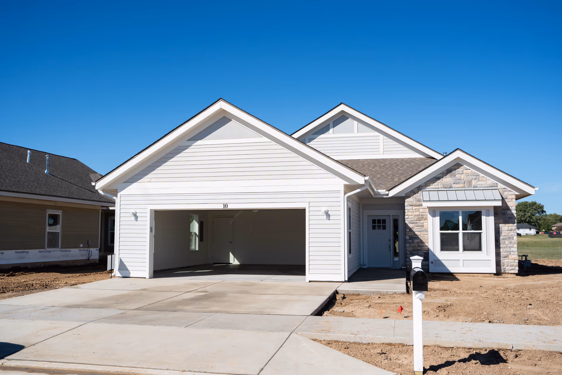 Exterior view of a newly constructed single-story house with white siding and stone accents, featuring an open two-car garage and a mailbox in front. The sky is clear and blue.