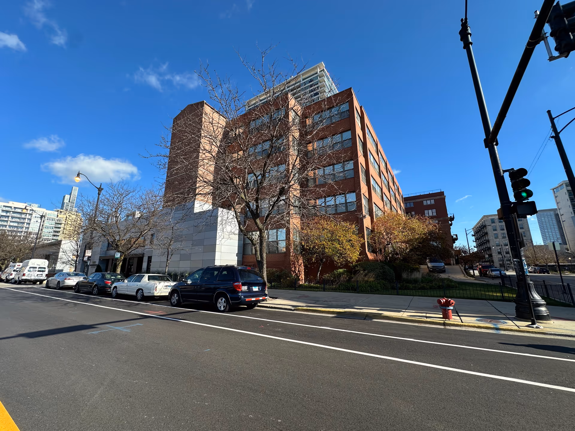 Street view of a multi-story brick building with large windows, leafless trees in front, several parked cars along the street, a fire hydrant on the sidewalk, and a traffic light showing green. The sky is clear and blue.