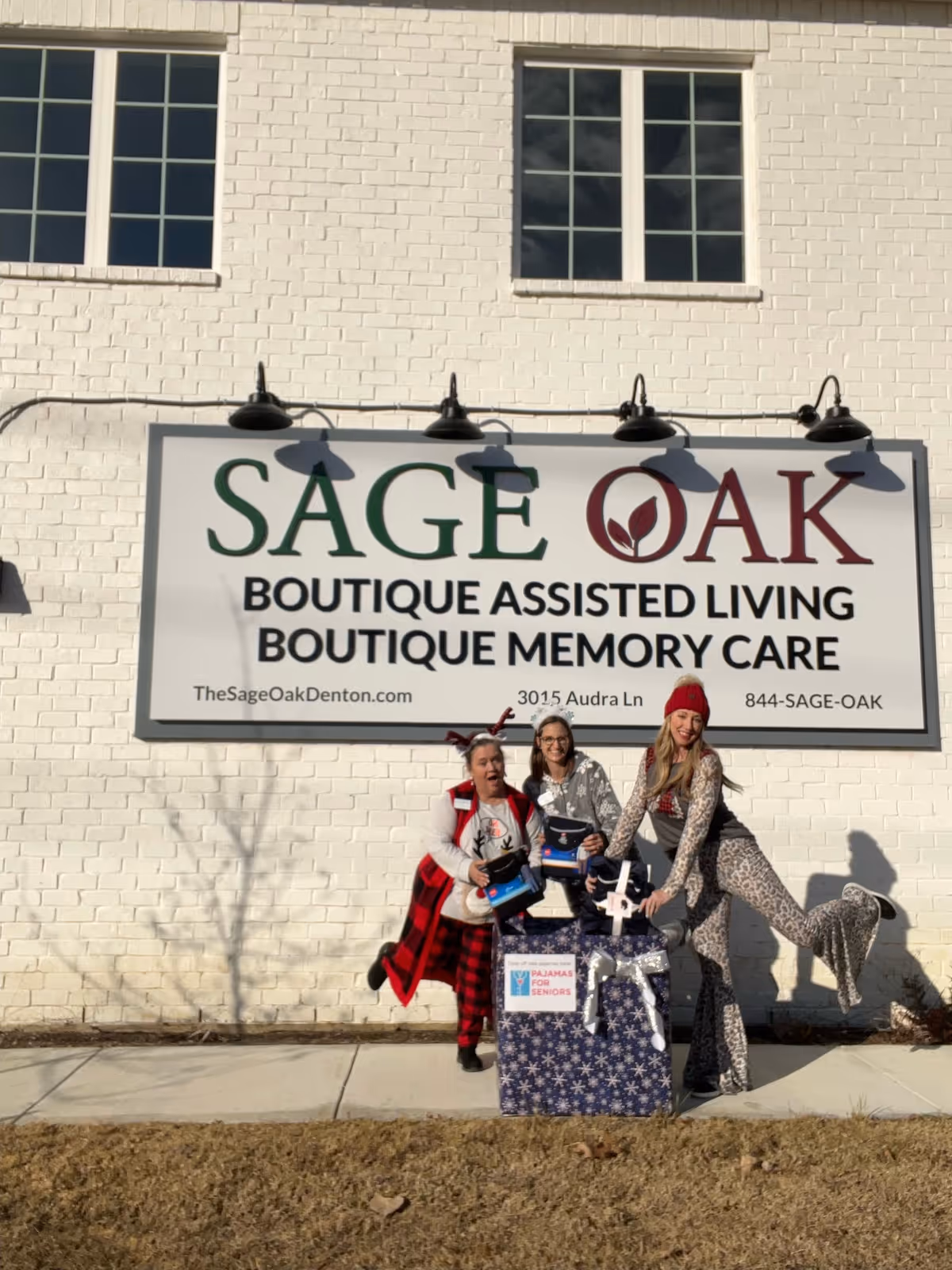 Three people pose in front of a large 'SAGE OAK Boutique Assisted Living Boutique Memory Care' sign on a white brick building.