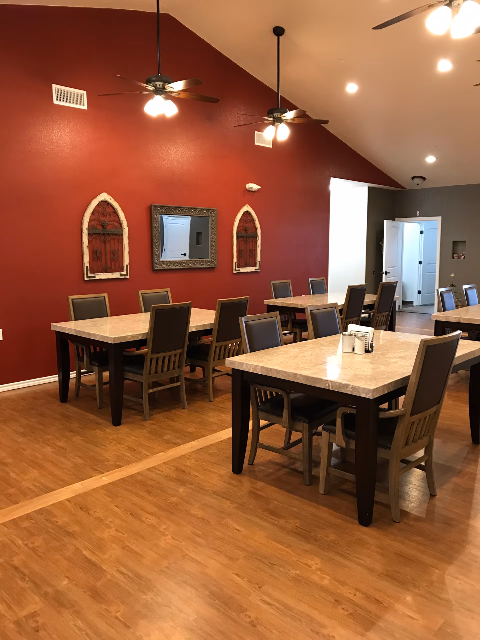 Spacious dining room with multiple rectangular tables and chairs, wood floors, a red accent wall with decorative mirrors, and ceiling fans.