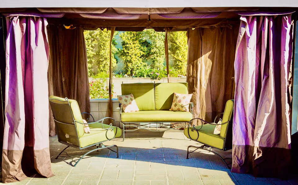 Outdoor seating area with green cushioned metal chairs and a loveseat under a canopy with brown curtains, surrounded by greenery and plants in the background.