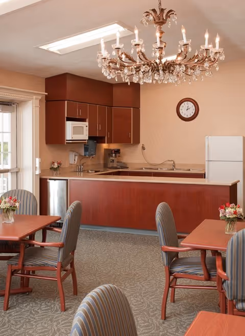 A dining area with wooden tables and striped cushioned chairs, each table decorated with a small vase of flowers. In the background, there is a kitchen area with wooden cabinets, a microwave, a coffee machine, a sink, and a refrigerator. A decorative chandelier hangs from the ceiling, and a clock is mounted on the wall.