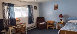 A simple, cozy bedroom with a single bed on the right side covered with a blue and white patterned blanket. Next to the bed is a small wooden nightstand with a red lamp and a small decorative item. There is a wicker chair and a brown recliner chair against a light blue wall. On the left side, there is a window with dark blue curtains and a small wooden desk with a chair, a vase with flowers, and some items on it. The floor is carpeted in dark blue.