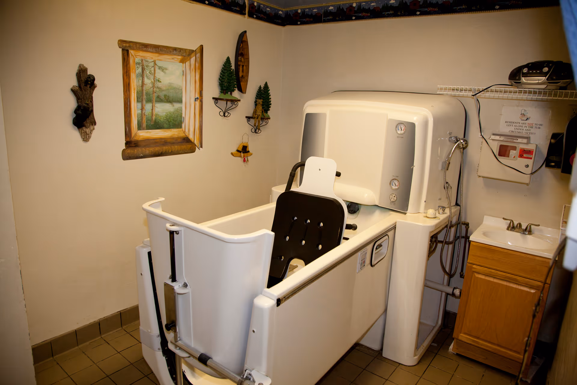 A small bathroom with a specialized bathing tub designed for assisted bathing, featuring a built-in seat and controls. The room has beige walls decorated with a painting of a forest scene and small wall-mounted shelves with decorative items. There is a sink with a wooden cabinet underneath and a small stereo on a wire shelf above it.