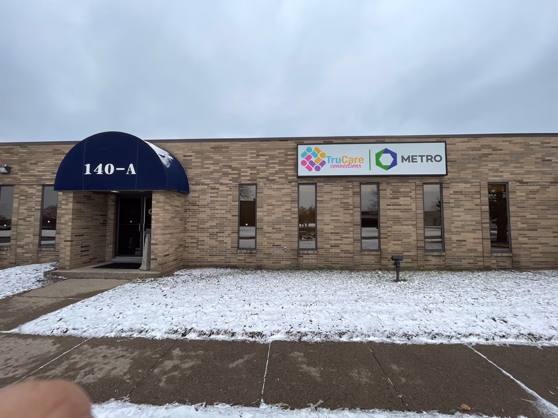 Brick commercial building with TruCare Connections sign and blue entrance awning labeled 140-A, snow-covered ground.