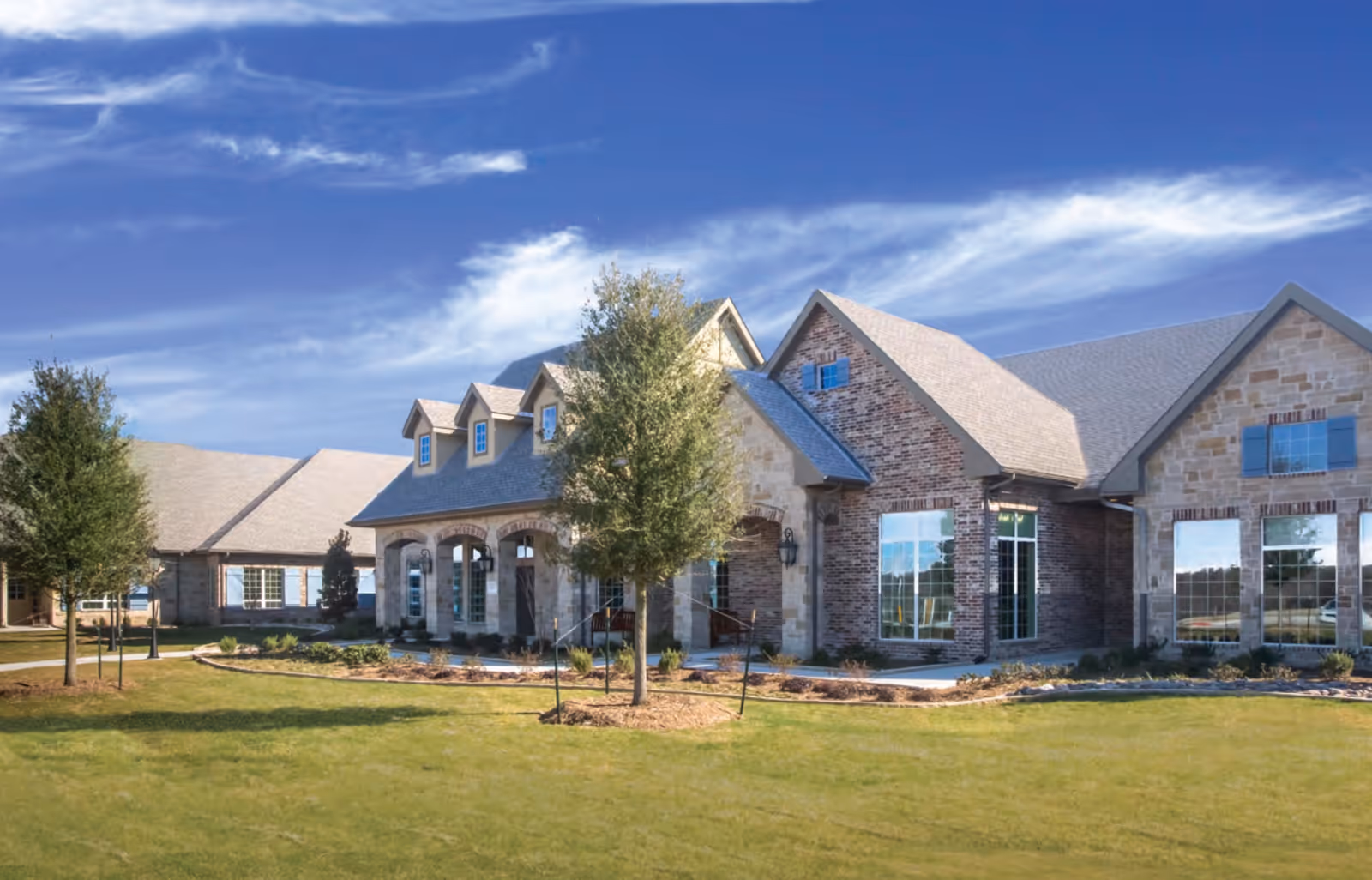 Front exterior of a single-story brick and stone senior living building with large windows, an arched entrance, and landscaped lawn under a blue sky.