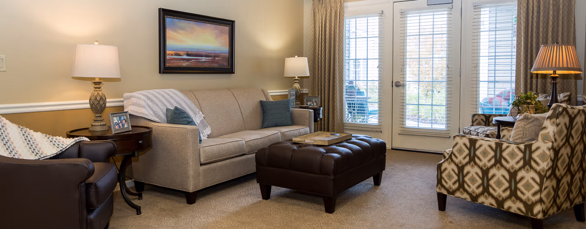 Cozy living room with beige sofa, patterned chair, ottoman, lamps, and glass doors