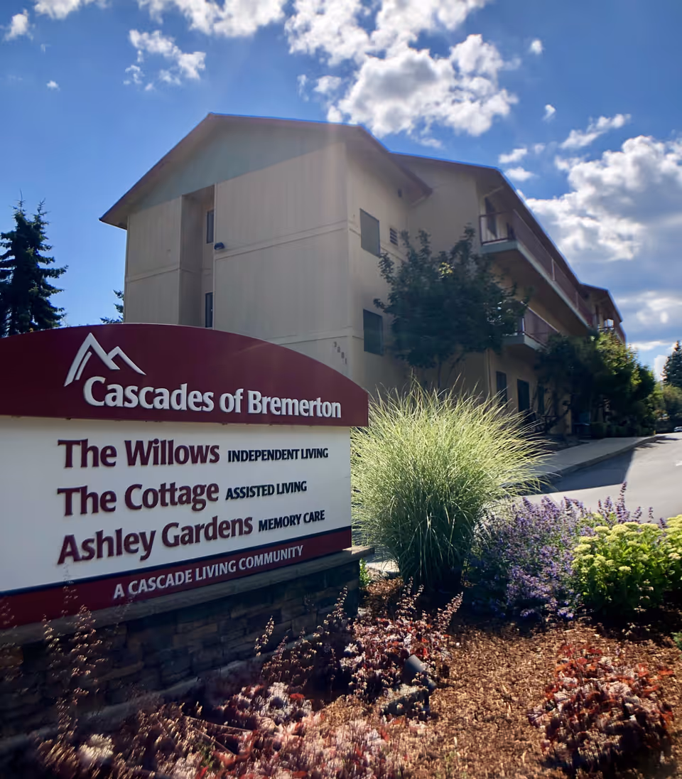 Outdoor view of the Cascades of Bremerton senior living facility sign with a beige multi-story building in the background, surrounded by landscaping including green and purple plants under a partly cloudy blue sky.