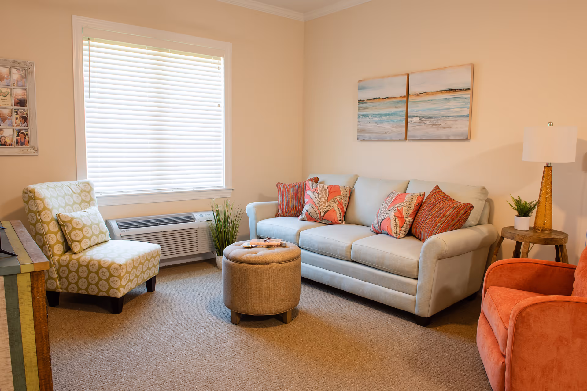 A cozy living room with a light blue sofa adorned with patterned throw pillows, a round beige ottoman in front, a green patterned armchair near a window with white blinds, and an orange armchair. A wooden side table with a lamp and a small plant is next to the sofa. Two paintings of a beach scene hang on the wall above the sofa.
