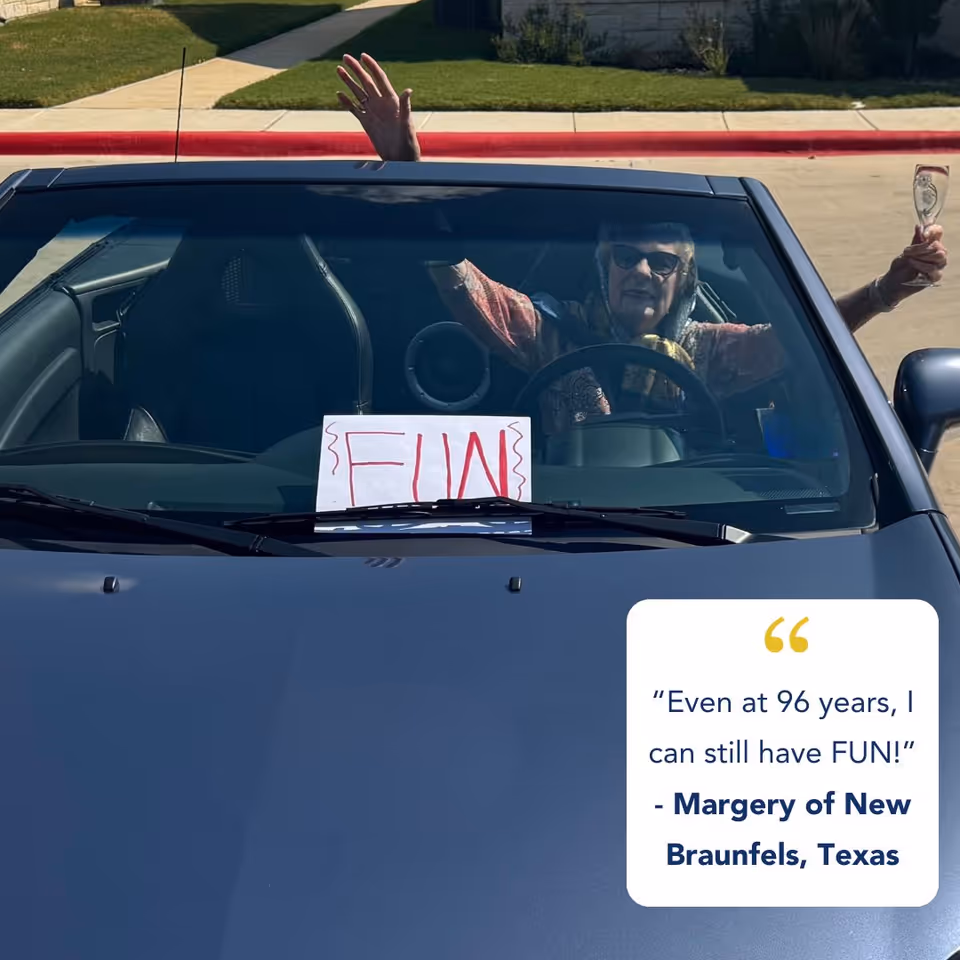 Elderly woman in a car waves from the driver's seat with a FUN sign on windshield