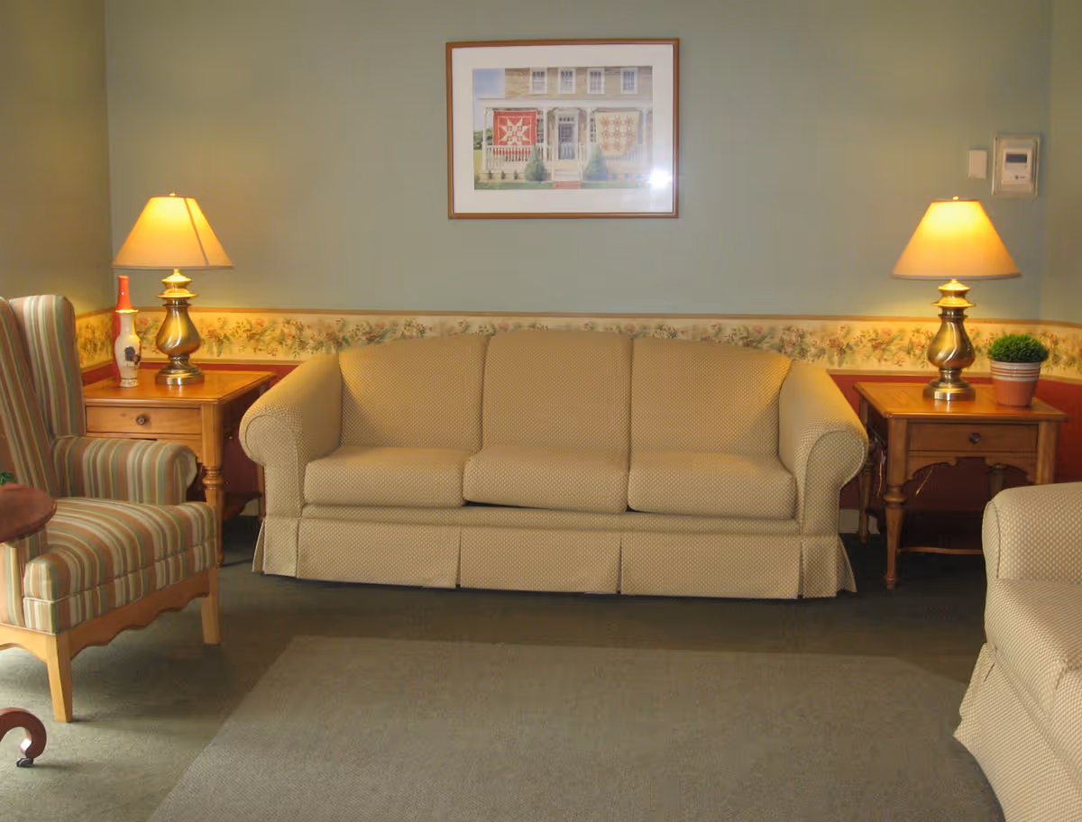 A cozy living room with a beige sofa centered between two wooden side tables topped with lamps, striped armchairs, and framed artwork on the wall.