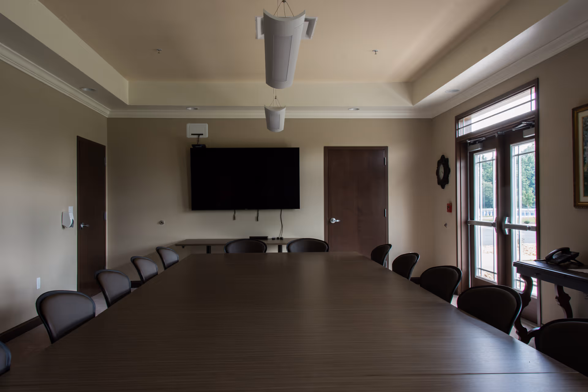 A long conference table surrounded by brown chairs in a well-lit meeting room with a TV.
