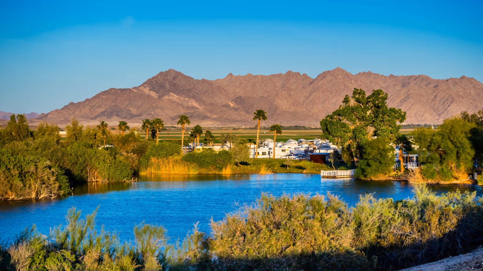 Blue river in foreground with trees and distant mountains behind an RV park