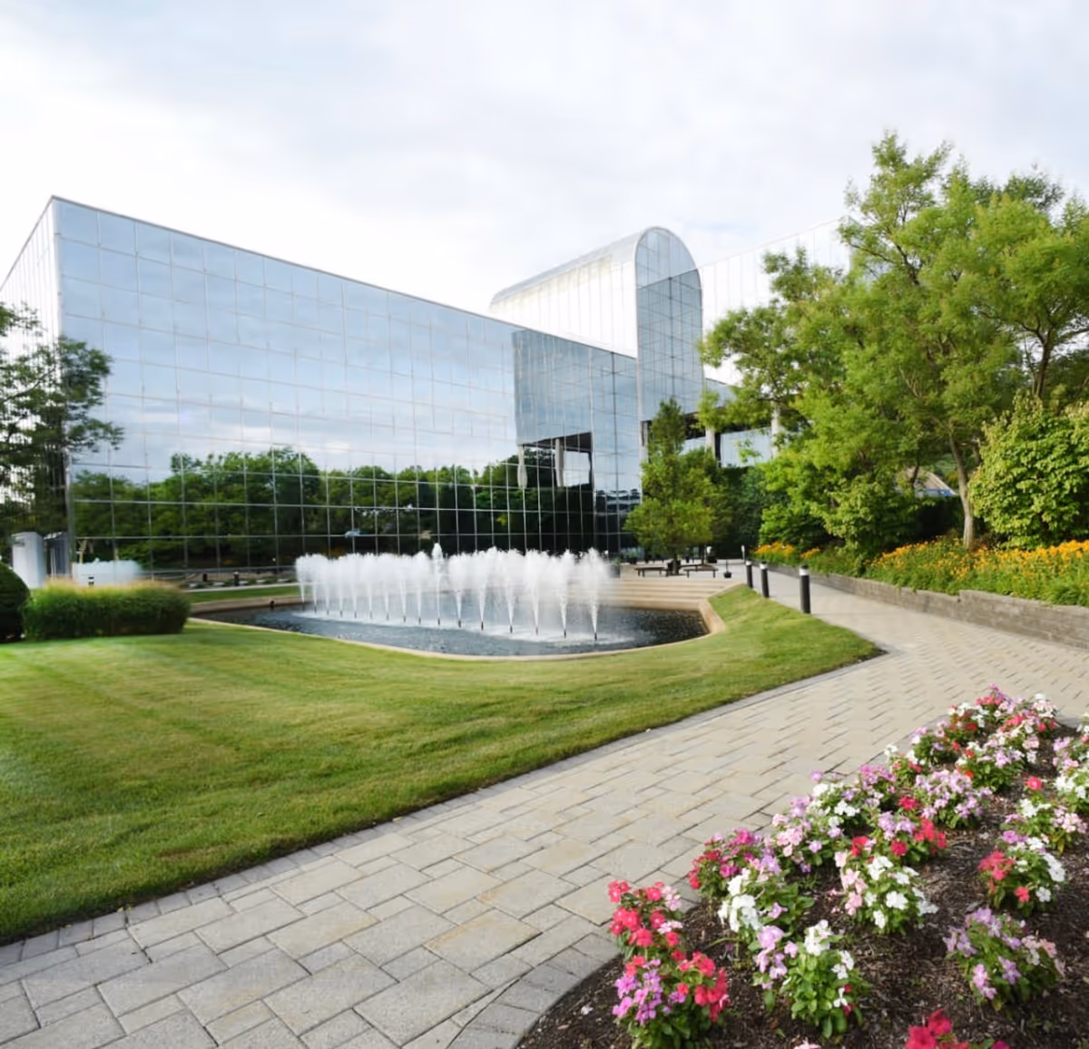 A modern glass building with a circular fountain, manicured lawn, and blooming flowers.