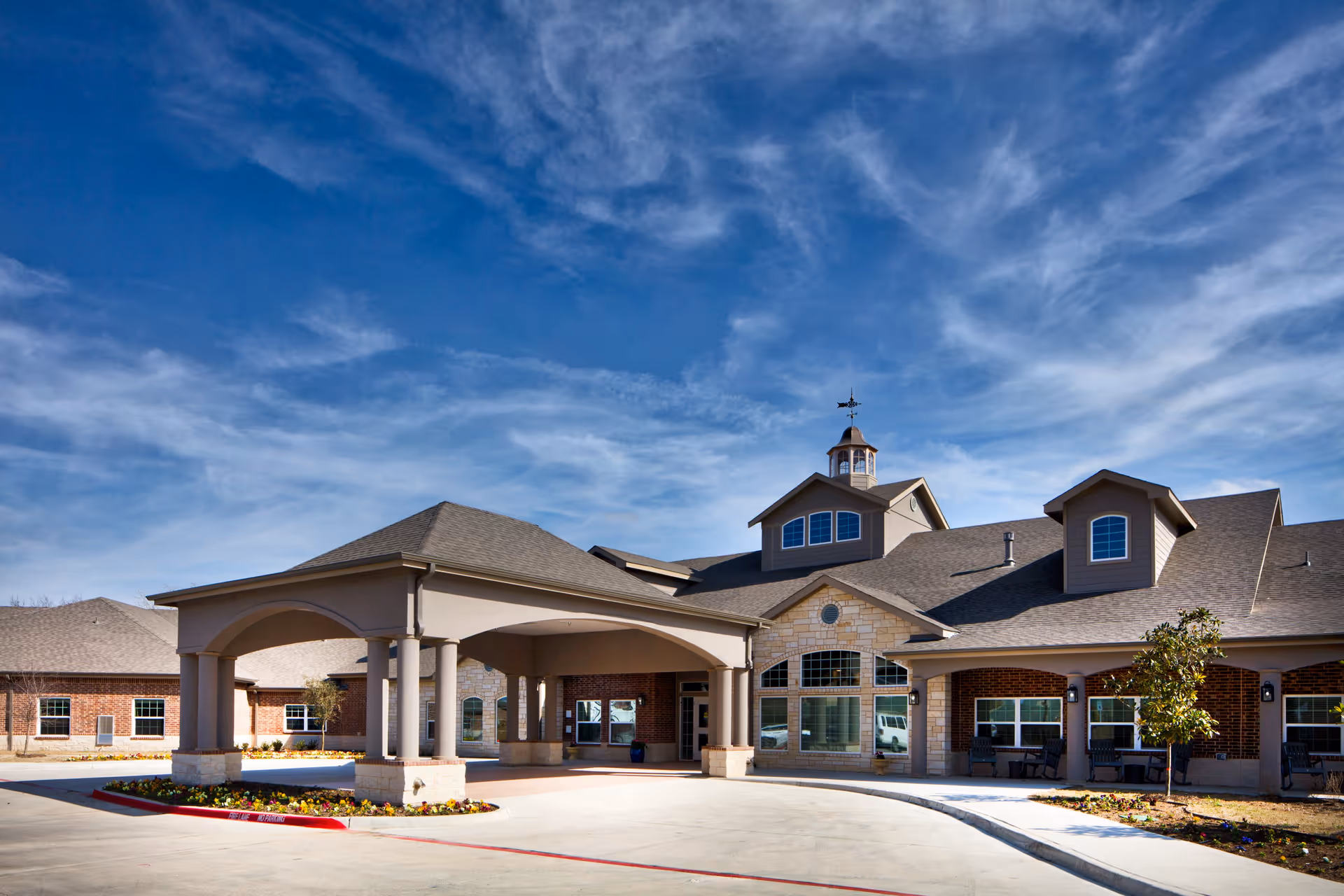 Front exterior view of Twin Rivers Assisted Living and Memory Care facility showing a large covered entrance with columns, brick and stone facade, multiple windows, and a clear blue sky with some clouds.