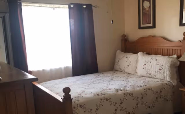 Sunlit bedroom with a wooden bed dressed in floral bedding, a dresser, and a curtained window.