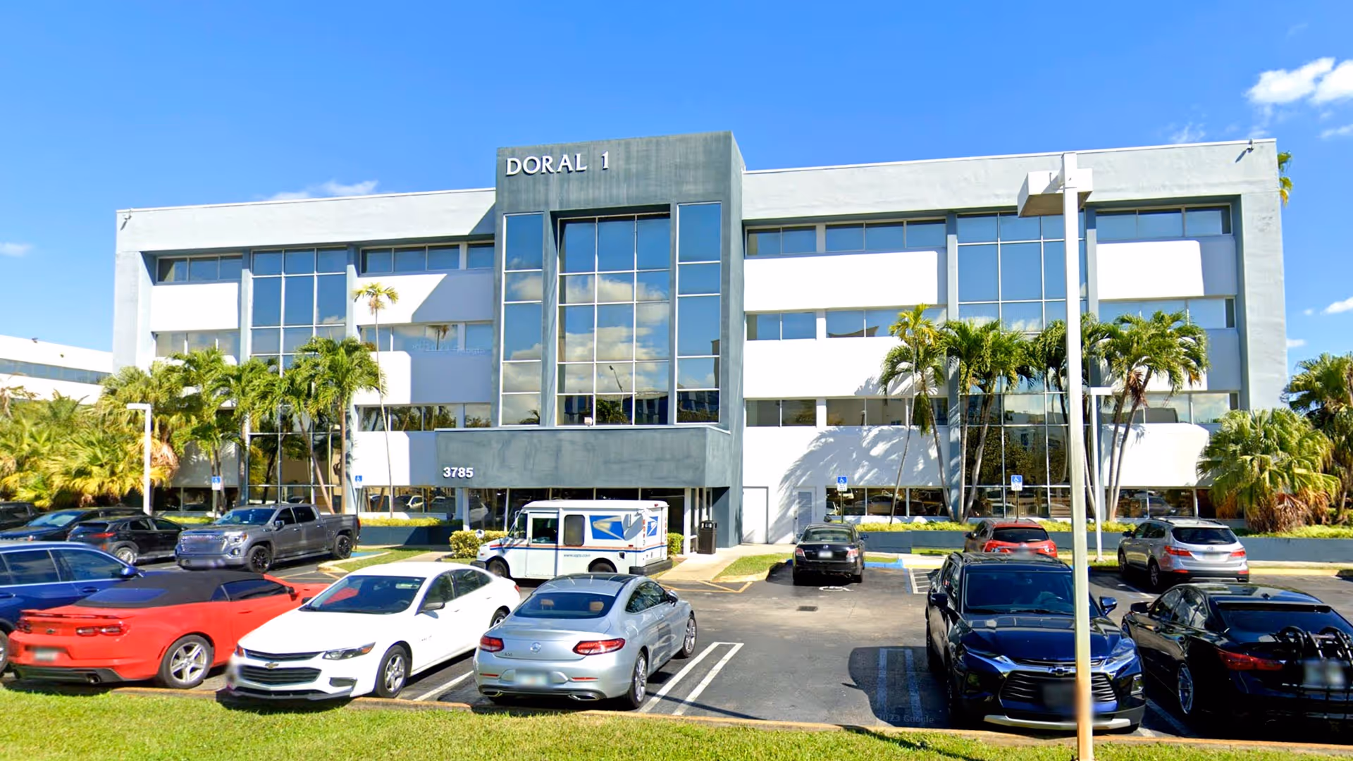 Exterior of a modern facility building with glass facade, palm trees, and parked cars.