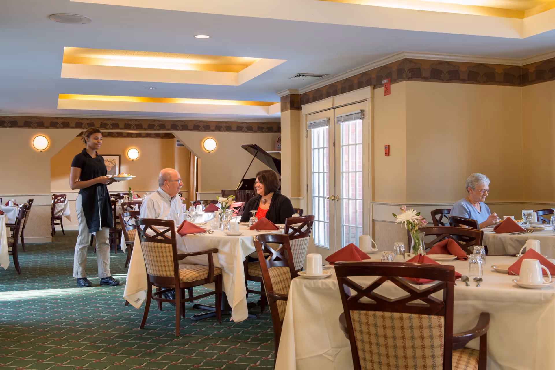 Dining room with several set tables, a server carrying plates, and residents seated and conversing.