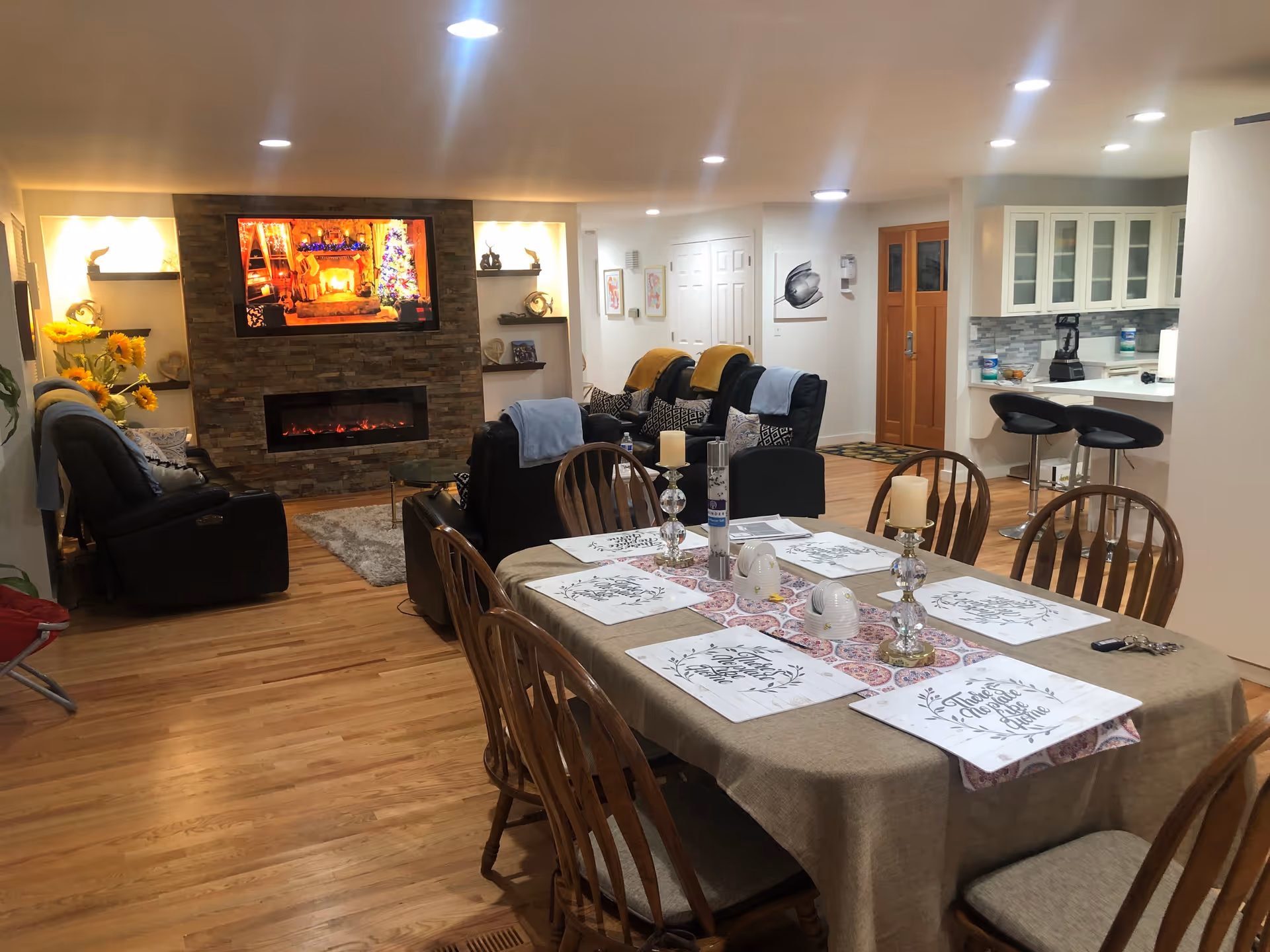 A cozy living and dining area with a wooden dining table set for six, decorated with placemats, candles, and a table runner. In the background, there are black recliner chairs with blankets, a stone fireplace with an electric fire and a TV mounted above it displaying a festive scene. The room has wooden flooring, recessed ceiling lights, and a kitchen area with bar stools and white cabinetry.