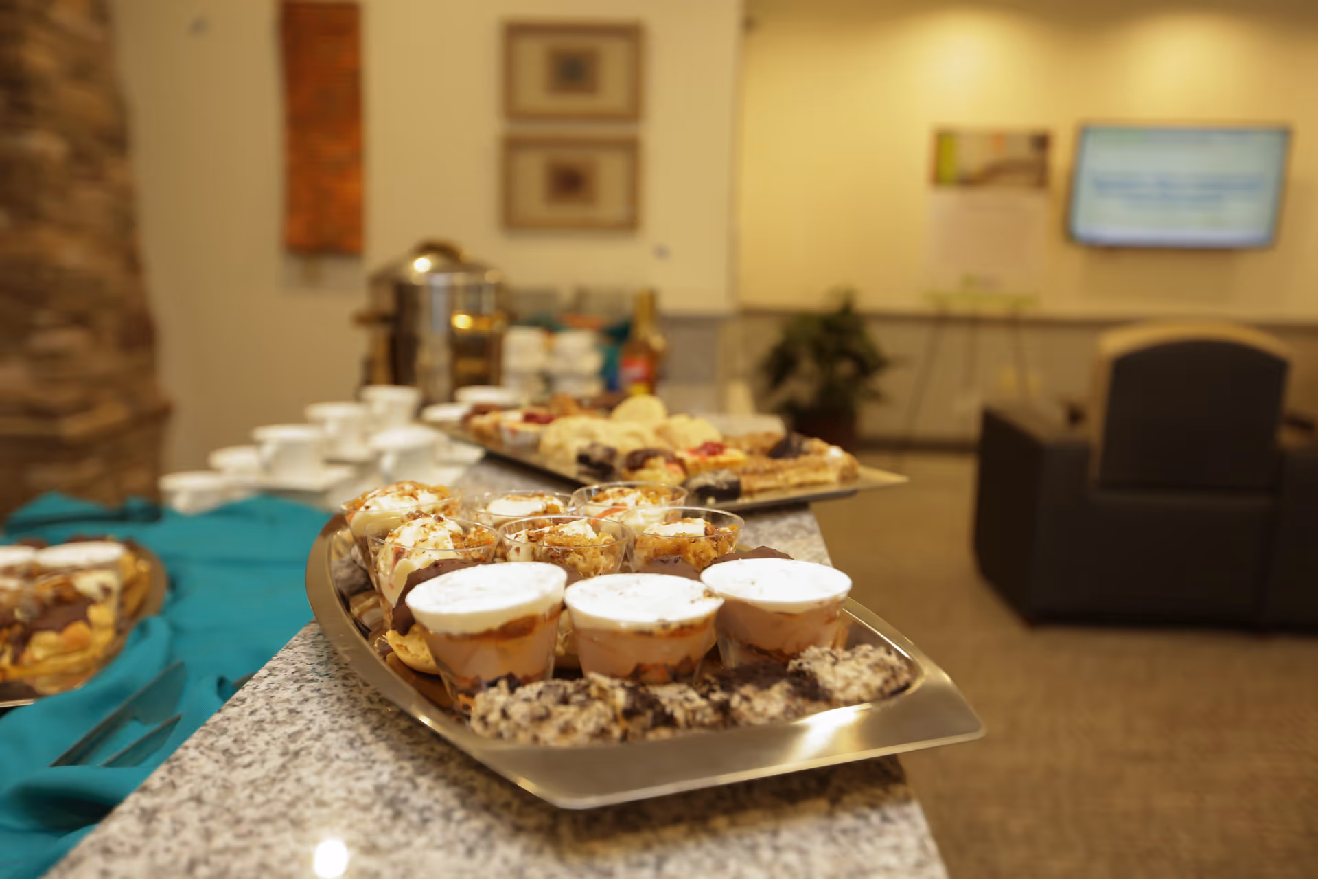 A close-up view of a buffet table with various desserts in small cups and plates, set on a granite countertop. In the background, there are white cups, a coffee urn, and a seating area with chairs and a TV mounted on the wall.