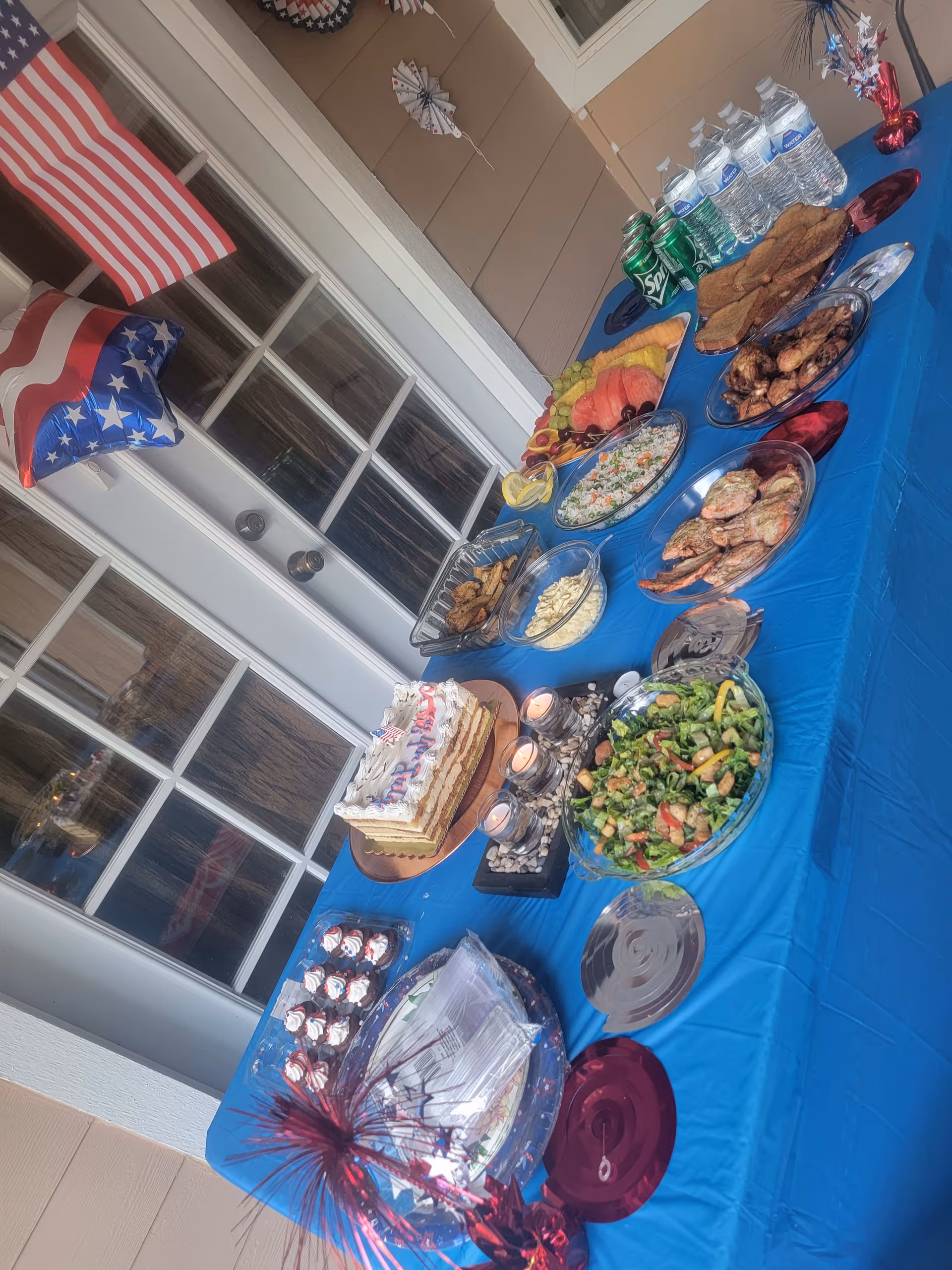 A table covered with a blue tablecloth displaying a variety of foods including a decorated cake, salad, fruit platter, grilled shrimp, chicken wings, cupcakes, and beverages such as bottled water and cans of soda. The table is set outdoors near a glass door with American flag-themed decorations including a flag and a star-shaped balloon.