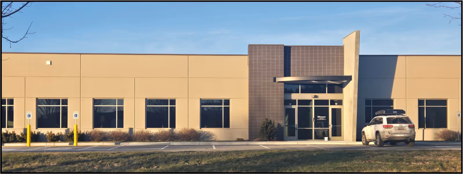 Exterior view of a single-story beige commercial building with a glass entrance and a parked SUV.