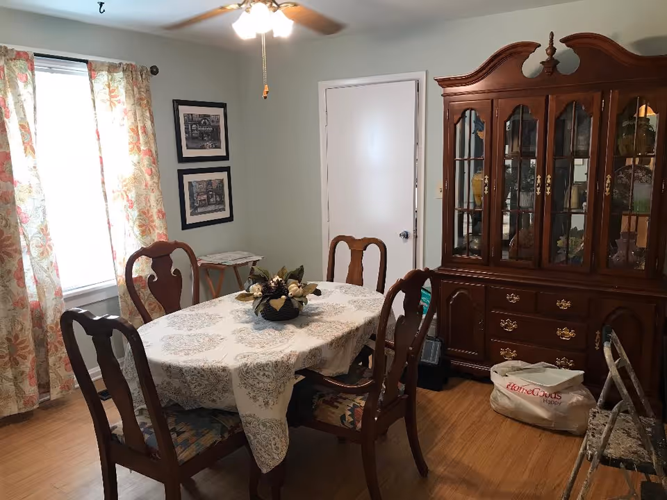 A traditional dining room with a floral tablecloth, wooden chairs, and a china cabinet.