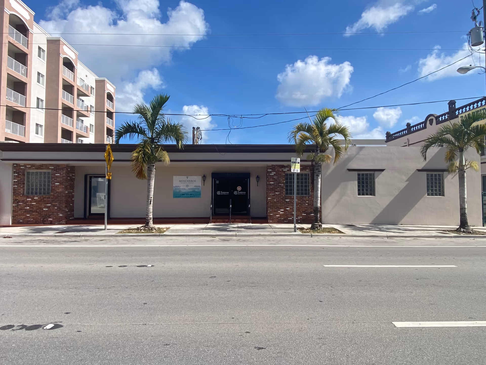 Single-story building storefront along a street with palm trees and signage.