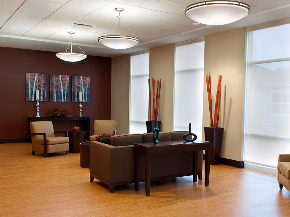A modern and spacious living room area with wooden flooring, beige and brown armchairs, a brown sofa, a dark wooden table with decorative vases, tall windows with white blinds, and wall art featuring a forest scene with red leaves. Two large ceiling lights illuminate the room.
