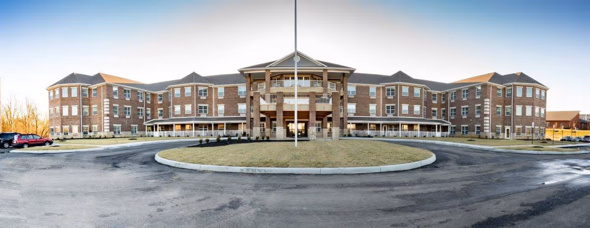 Front exterior view of a large three-story brick senior living facility building with multiple windows, a central entrance with columns, and a circular driveway with a grassy island in the middle.