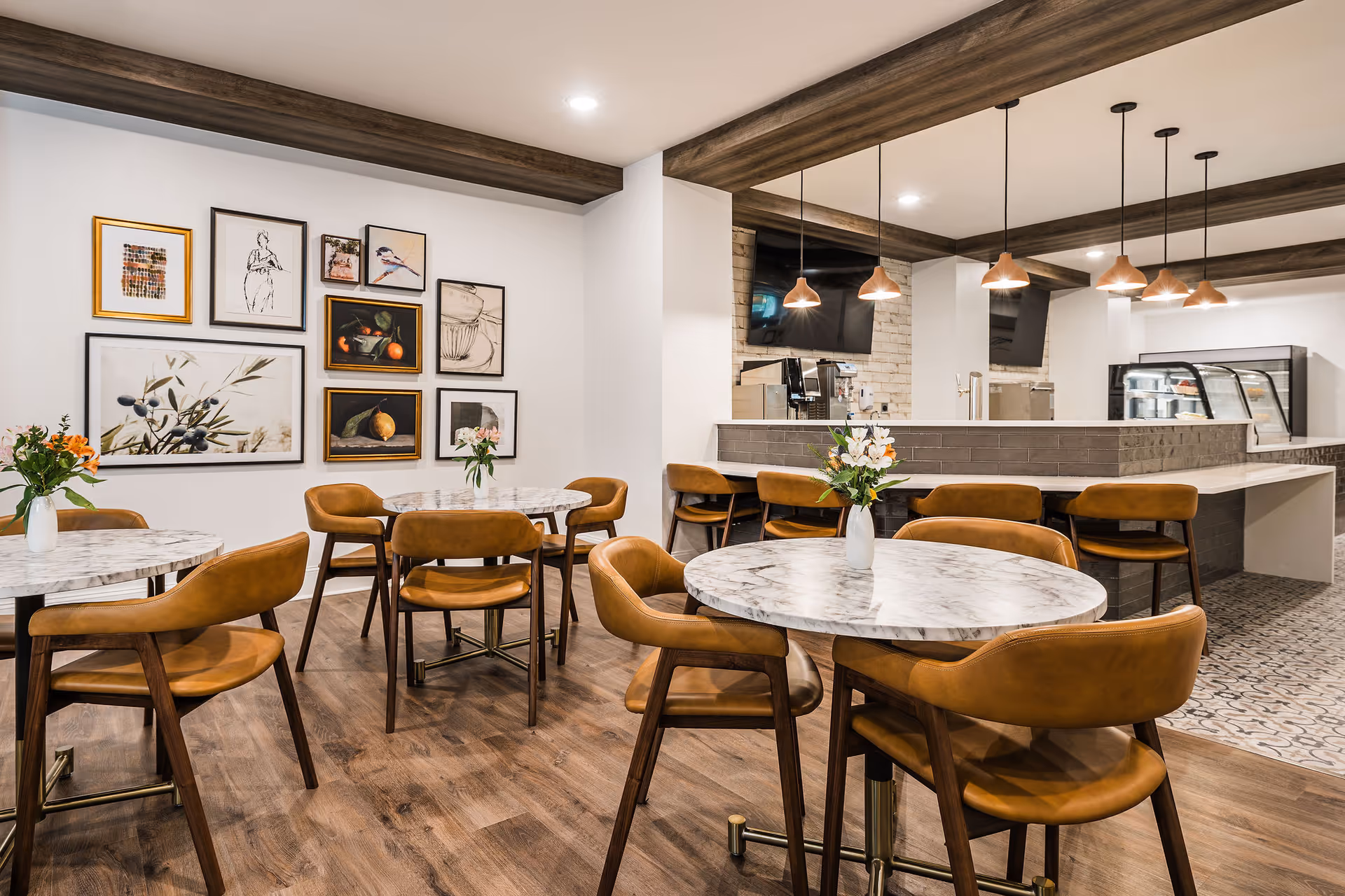 A modern dining area with round marble tables and brown leather chairs. The room features wooden flooring, a wall decorated with framed artwork, and a counter area with pendant lights hanging from the ceiling. There are flower vases on the tables and a display refrigerator behind the counter.